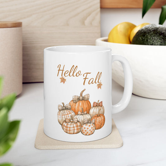 White ceramic coffee mug with a watercolor illustration of several orange and white pumpkins, each with a plaid or patterned bow, with "Hello Fall" text and fall leaves above.