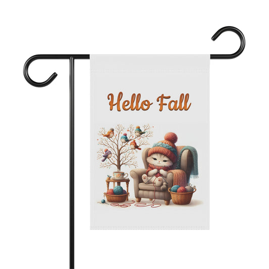 A 'Hello Fall' garden flag on a metal stand, featuring a cute, cozy cat knitting in an armchair with a tree and birds.