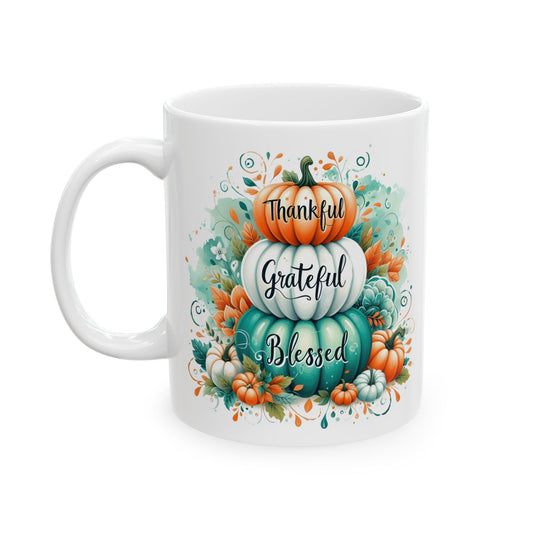 White ceramic coffee mug with a watercolor illustration of three stacked pumpkins (orange, white, teal) with the words "Thankful Grateful Blessed" written on them, surrounded by fall leaves and small pumpkins.