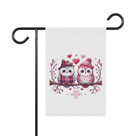 Happy Valentine's Day garden flag with a charming illustration of two cartoon owls wearing winter hats and scarves, sitting on a branch surrounded by hearts. The flag is hanging on a black metal holder.

