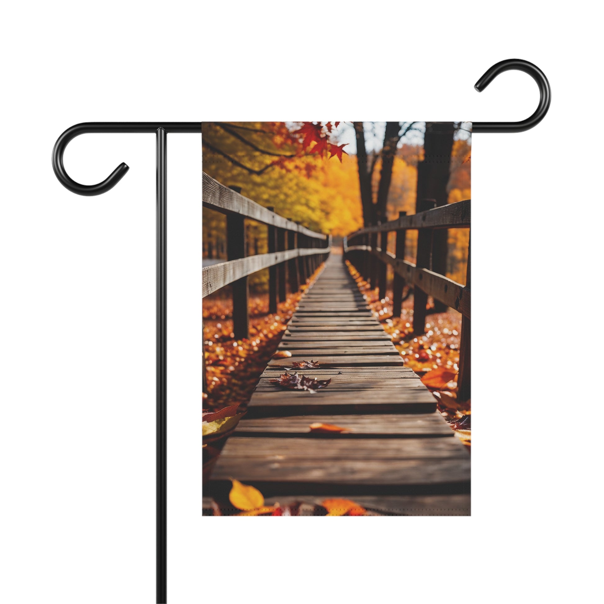 Garden flag depicting a close-up, perspective view of a wooden bridge covered in fallen orange and red autumn leaves, leading into a forest with vibrant fall foliage. 