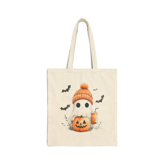 A cream-colored canvas tote bag featuring an illustration of a cute ghost wearing an orange beanie, holding a jack-o'-lantern and a cup with a straw. Bats are flying in the background.