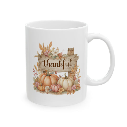 White ceramic coffee mug printed with brown Thankful script on a wooden sign surrounded by orange, pink, and white pumpkins, wheat, and fall blossoms