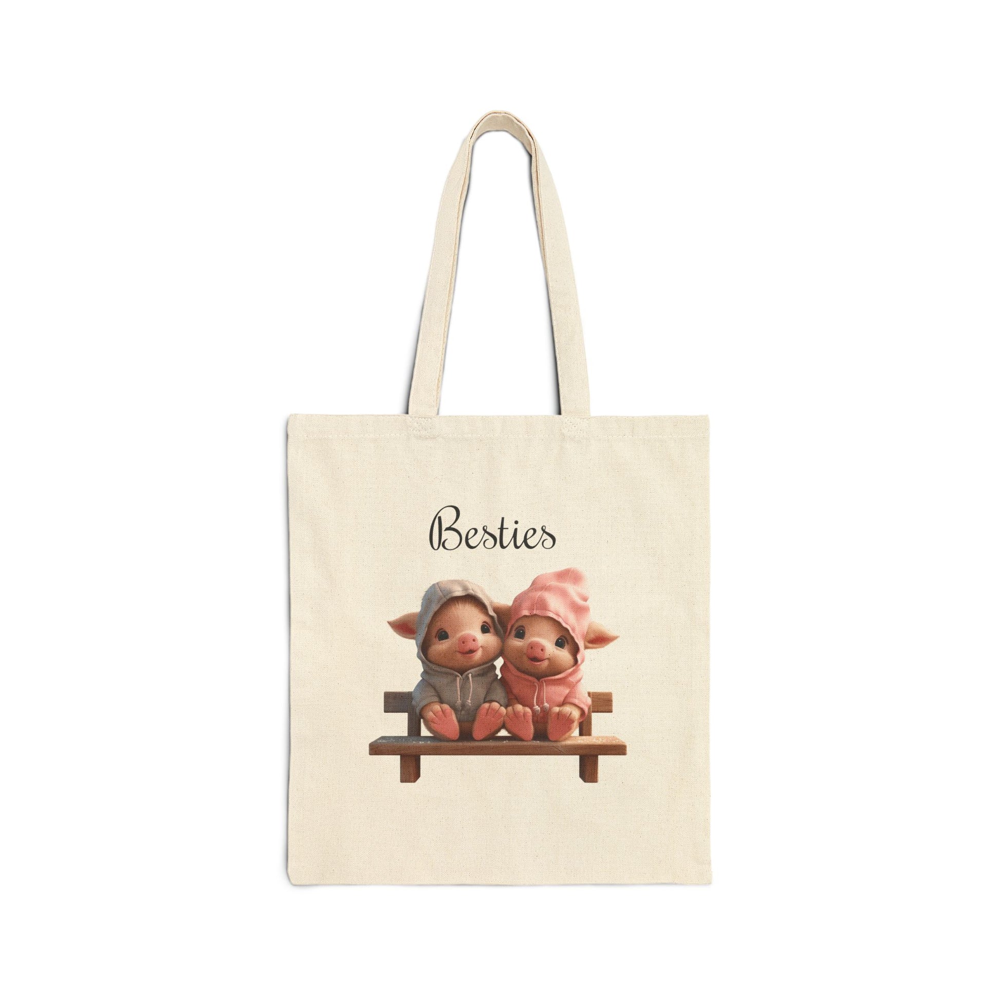 A cream-colored 100% cotton canvas tote bag featuring an illustration of two small, cute piglets in hoodies sitting on a wooden bench with the word "Besties" above them.