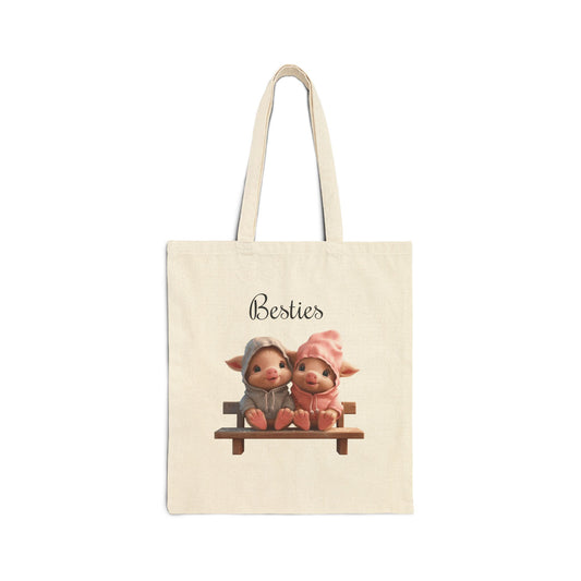  A cream-colored 100% cotton canvas tote bag featuring an illustration of two small, cute piglets in hoodies sitting on a wooden bench with the word "Besties" above them.