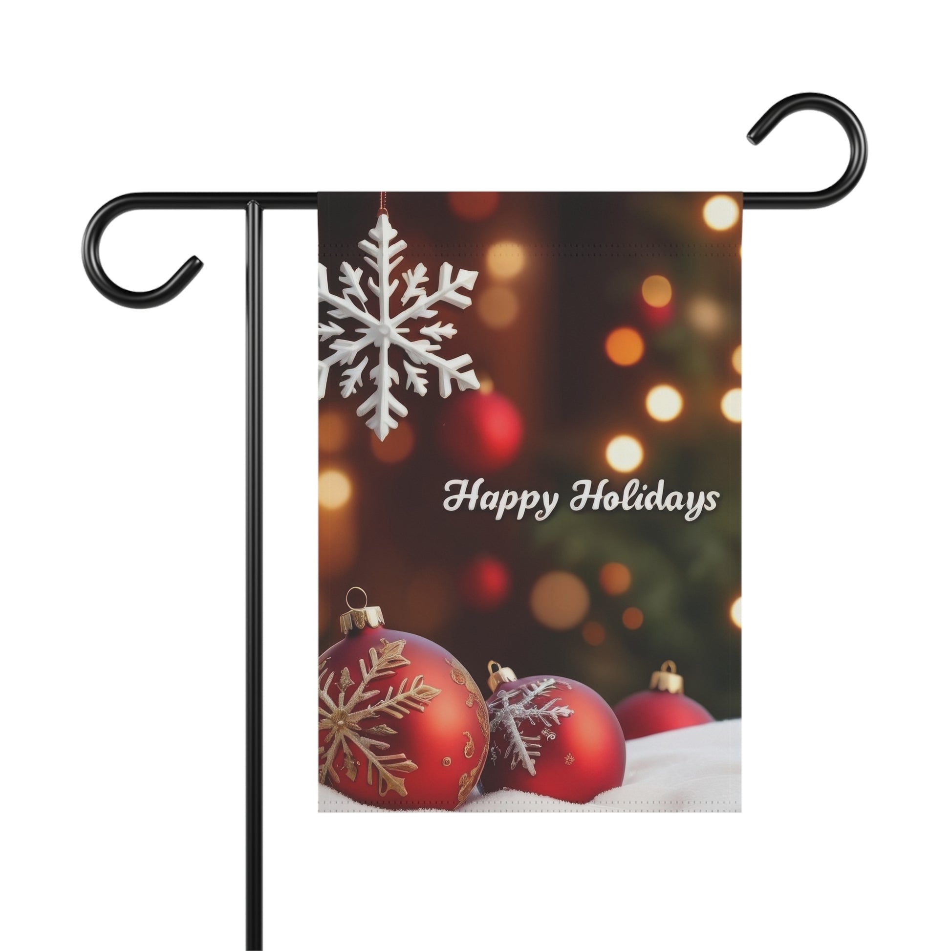 Garden flag with "Happy Holidays" script, featuring a white snowflake hanging at the top, and three red Christmas ornaments with white designs resting on a white surface at the bottom, with blurred festive lights in the background.

