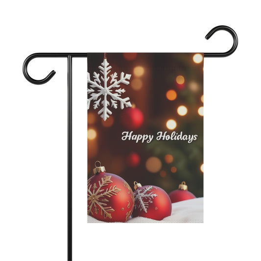 Garden flag with "Happy Holidays" script, featuring a white snowflake hanging at the top, and three red Christmas ornaments with white designs resting on a white surface at the bottom, with blurred festive lights in the background.

