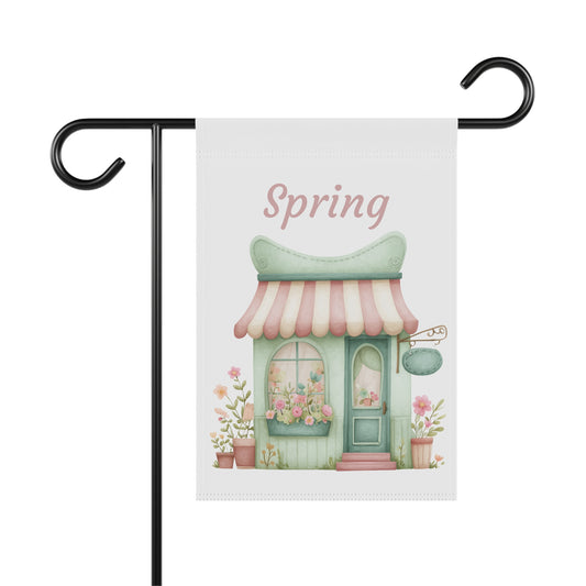 Outdoor garden flag featuring a watercolor illustration of a pastel green flower shop cottage beneath pink script text reading Spring.