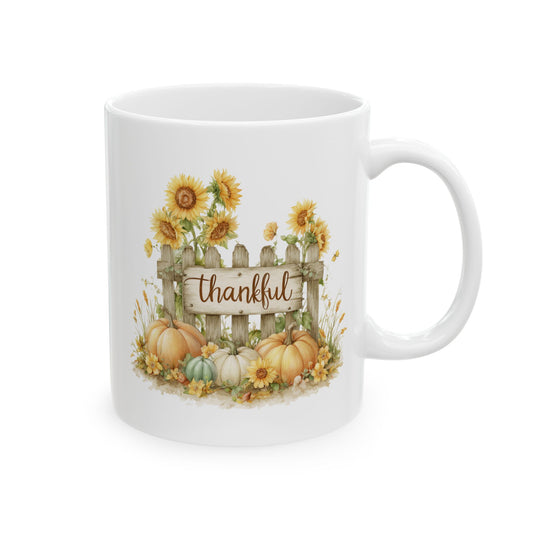 White ceramic coffee mug printed with brown Thankful script on a wooden fence sign surrounded by bright sunflowers and colorful pumpkins