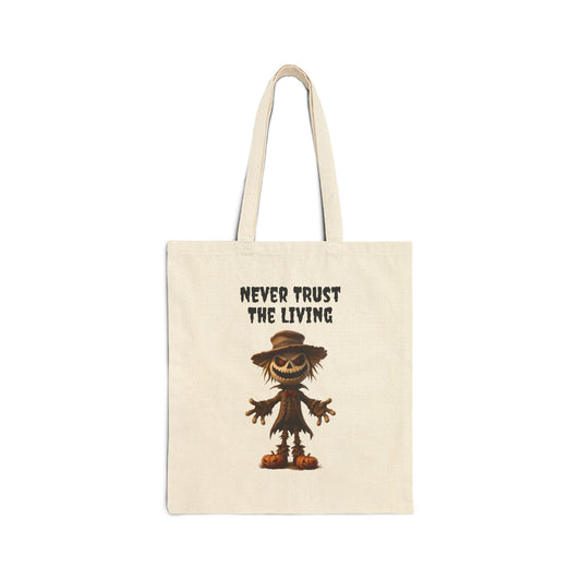 A cream-colored 100% cotton canvas tote bag featuring an illustration of a spooky, grinning scarecrow with a stitched mouth and a hat. The text "NEVER TRUST THE LIVING" is printed in a distressed font above the scarecrow.
