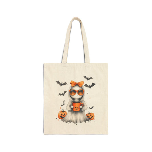 A cream-colored canvas tote bag featuring a cute ghost illustration wearing orange sunglasses and an orange bow, holding a pumpkin mug. Bats are flying in the background, and pumpkins are on the ground.