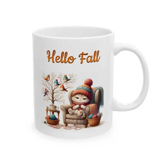 White ceramic coffee mug with a cozy illustration of a cute cat knitting in an armchair, with a tree full of birds nearby, and the words "Hello Fall" in orange script text above the scene.

