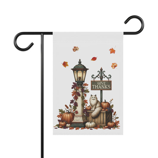 Whimsical Thanksgiving garden flag featuring a glowing vintage lamppost wrapped in fall leaves, a Give Thanks sign, and a fluffy white long-haired cat sitting on a wooden crate surrounded by pumpkins.