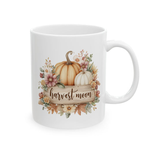 White ceramic coffee mug printed with brown Harvest Moon script on a rustic wood plaque below orange and white pumpkins framed by fall flowers