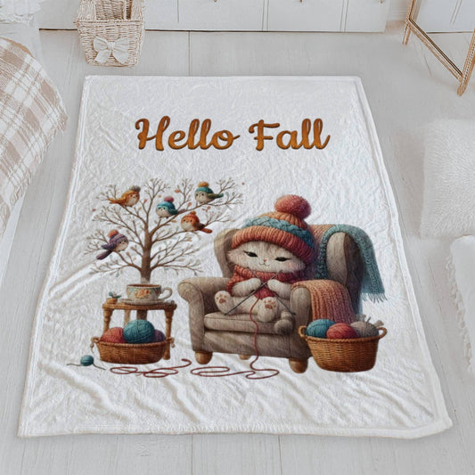 White velveteen blanket with a cozy autumn illustration of a cute cat knitting in an armchair, with birds in hats on a bare tree, and the words "Hello Fall" in orange script text above the scene.

